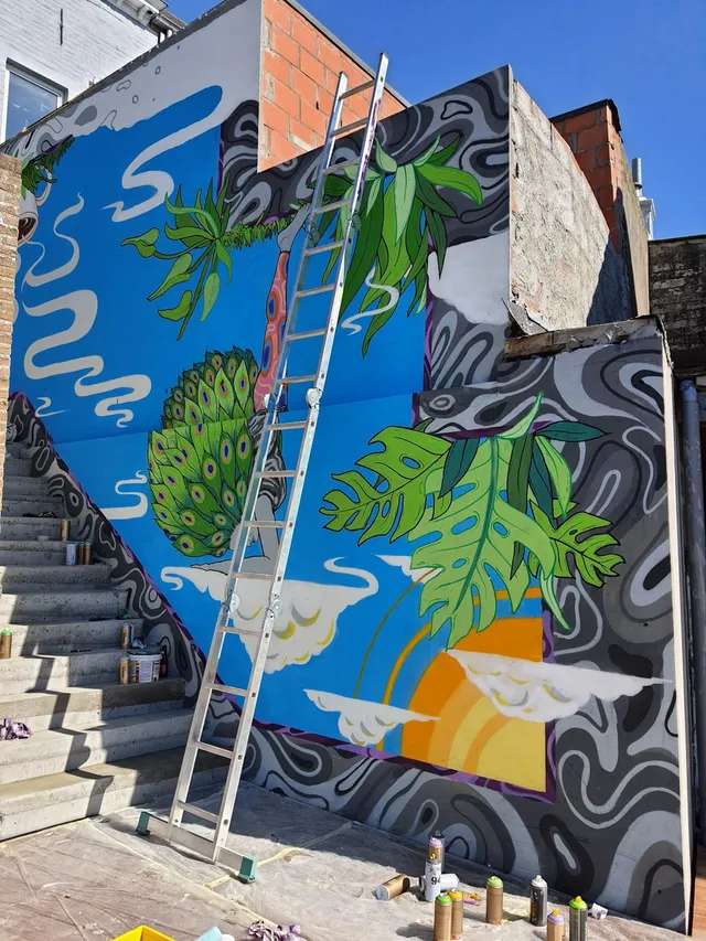 Mural in progress featuring a peacock, monstera leaves, and clouds on a blue background with wavy grey borders