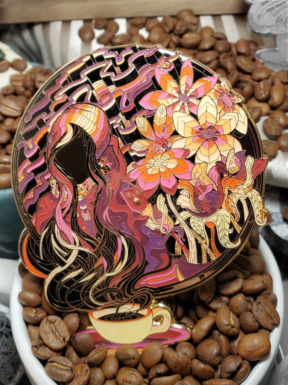 Psychedelic enamel pin of a hooded figure, flowing hair, coffee cup and bright flowers, resting on coffee beans