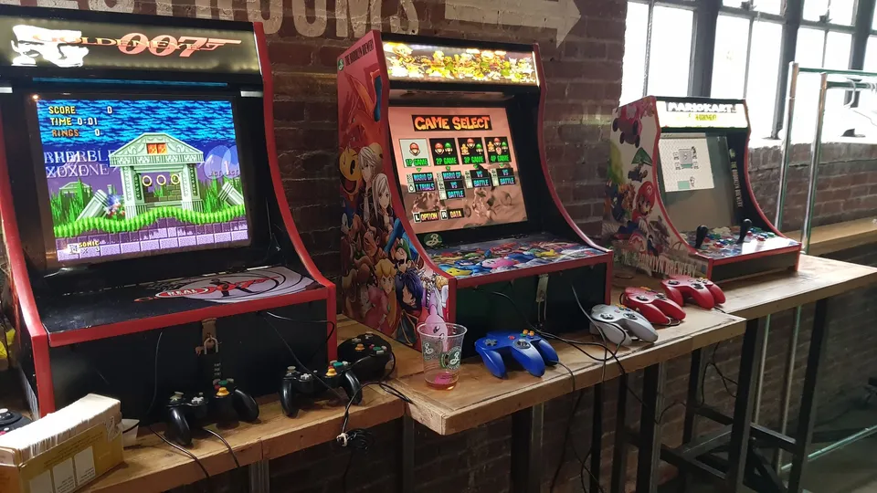 Three arcade machines with colorful artwork and game screens displayed on wooden tables against a brick wall