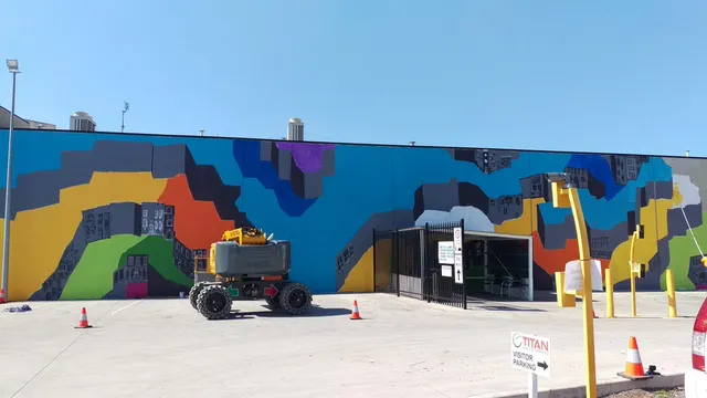 Large outdoor mural in progress featuring bright abstract color blocks and grey cityscapes, behind a parked boom lift