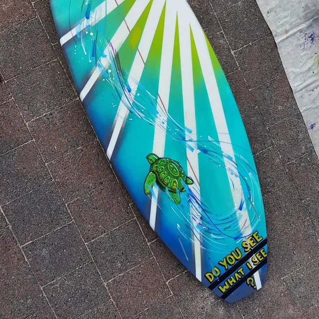 Painted surfboard with a green sea turtle, blue and green sunburst rays, and text DO YOU SEE WHAT I SEE?