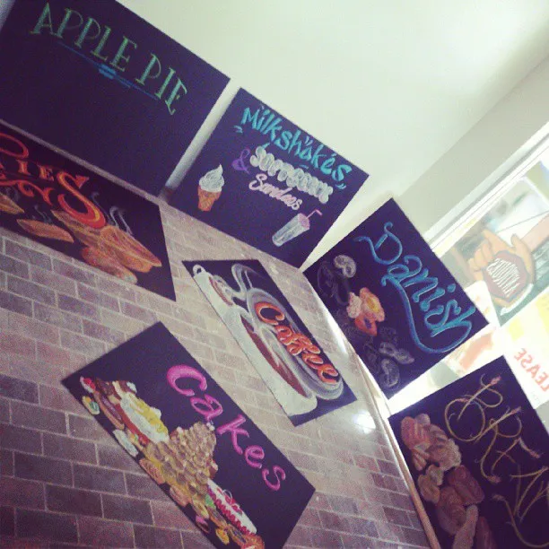 Hand-painted chalkboard signs on a brick wall displaying colorful lettering and illustrations of various cafe items