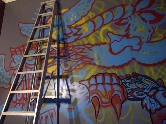 In-progress spray paint mural showing red, blue and yellow psychedelic wings and a claw, behind a metal ladder