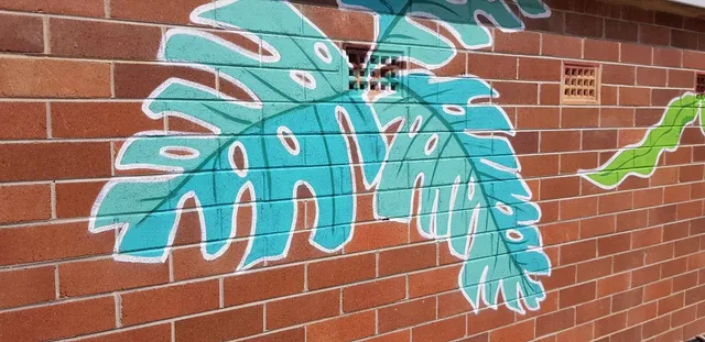 Mural of large teal monstera leaves with bold white outlines painted on a red brick wall