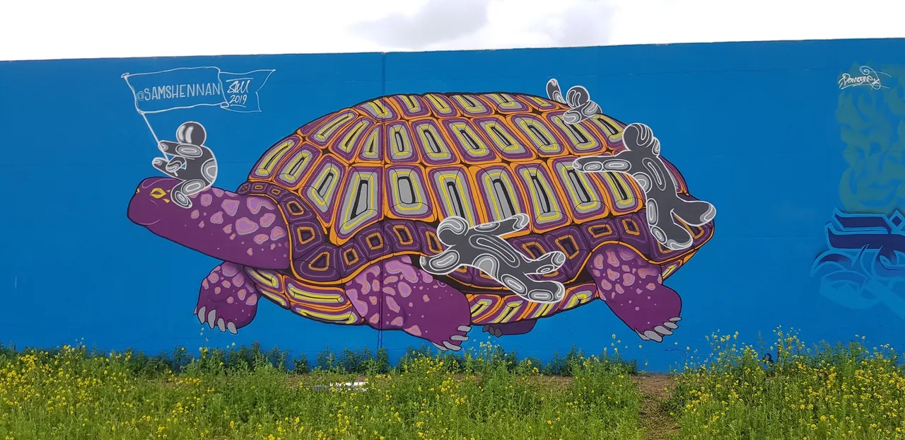 Mural on a blue wall of a giant purple tortoise with a geometric orange shell, ridden by small stylized grey figures