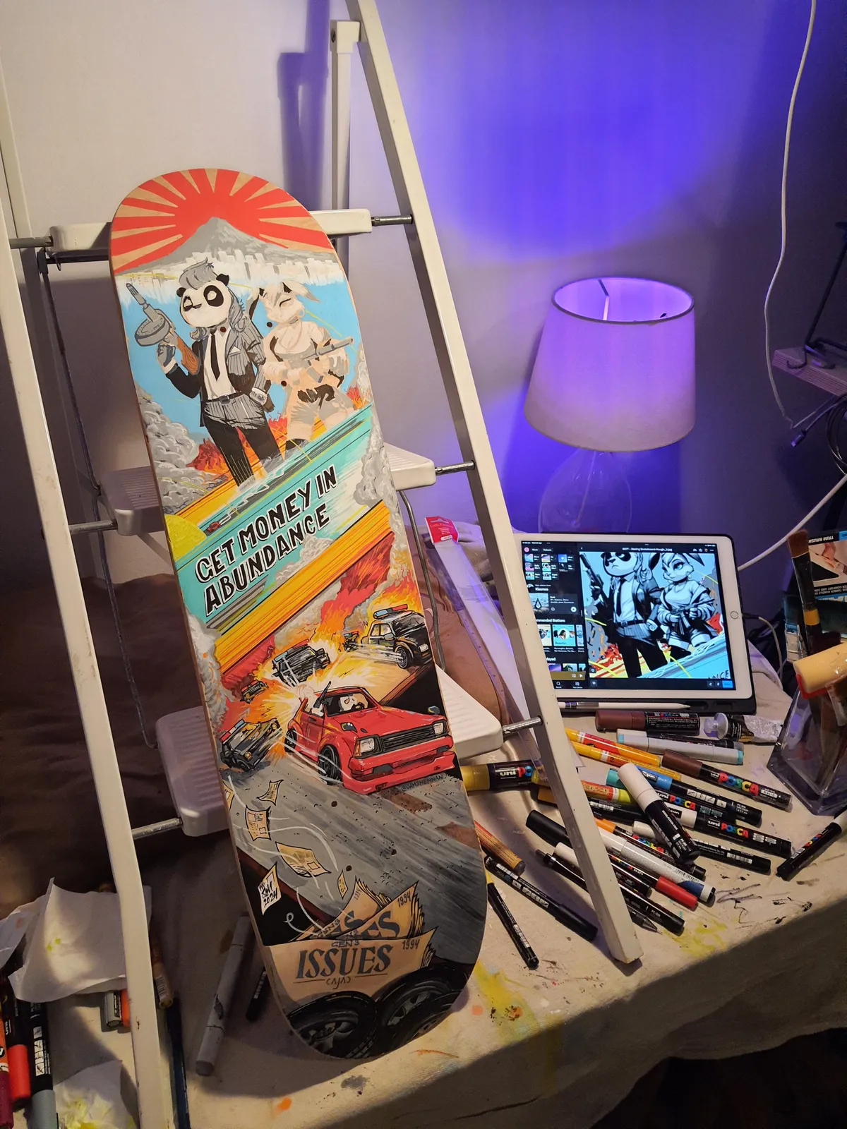 Hand-painted skateboard on a desk featuring a panda gangster, car chase, and text GET MONEY IN ABUNDANCE