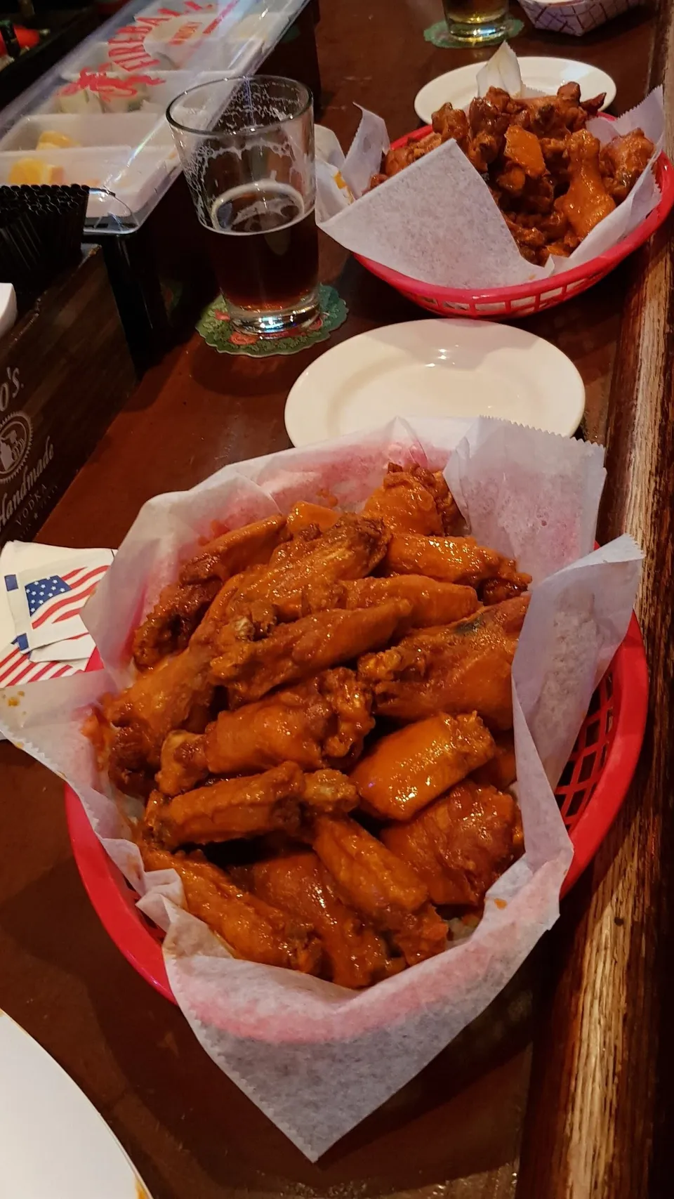 Two red baskets filled with buffalo wings on parchment paper, a glass of dark beer, and condiments on a wooden table
