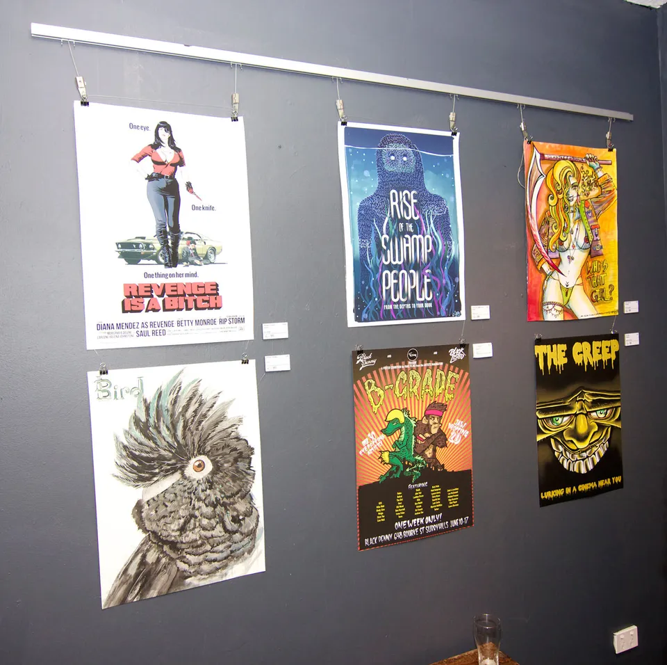 Six illustrated B-movie posters hanging from clips on a dark gallery wall