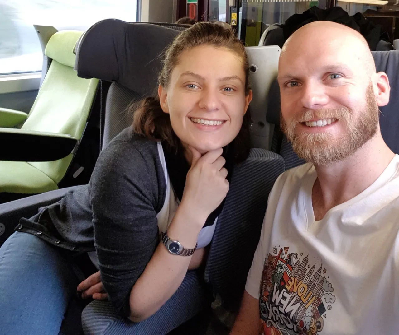 Two people smiling in a train seat, one wearing a graphic t-shirt with bold colorful artwork