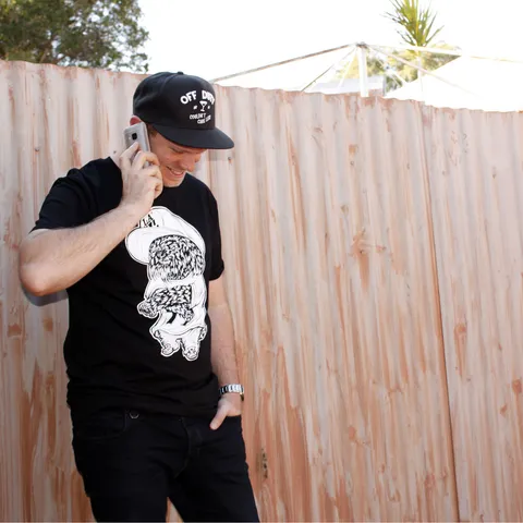 Man on a phone wearing a black t-shirt printed with a white bold line illustration of a dog in a hip hop cap