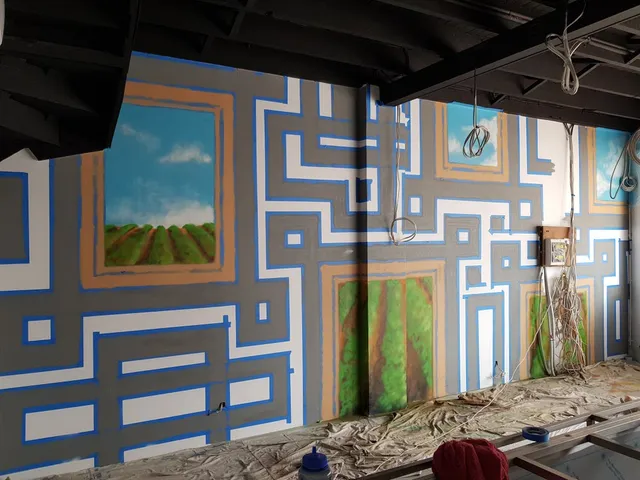 In-progress wall mural with blue painter's tape creating geometric patterns around painted windows showing fields and sky