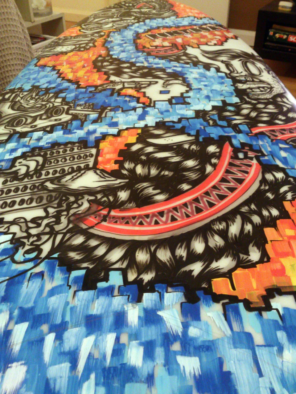 Painted surfboard detail showing bold black linework of a hairy totemic face amidst blue and orange pixelated blocks