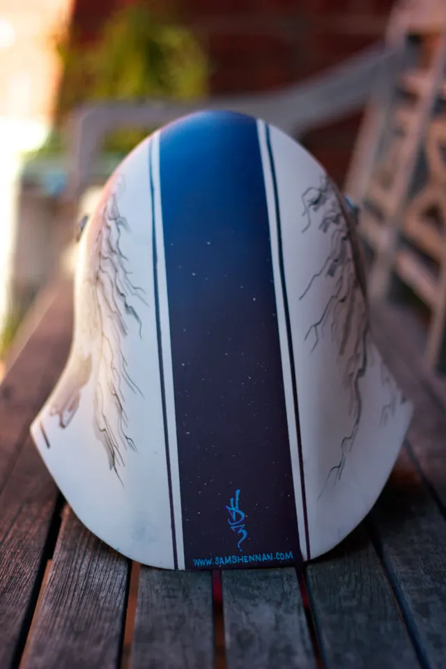 Back of a hand-painted white helmet with a dark speckled center stripe and symmetrical flowing line art on the sides