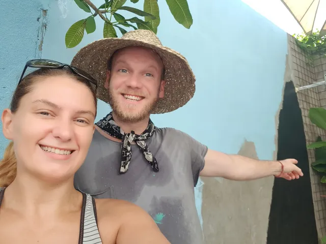 Woman taking a selfie with a smiling man in a straw hat pointing at a blue mural in progress