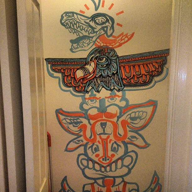 Wall mural in bold blue and orange line work depicting a psychedelic totem pole of stacked skulls, birds, and faces