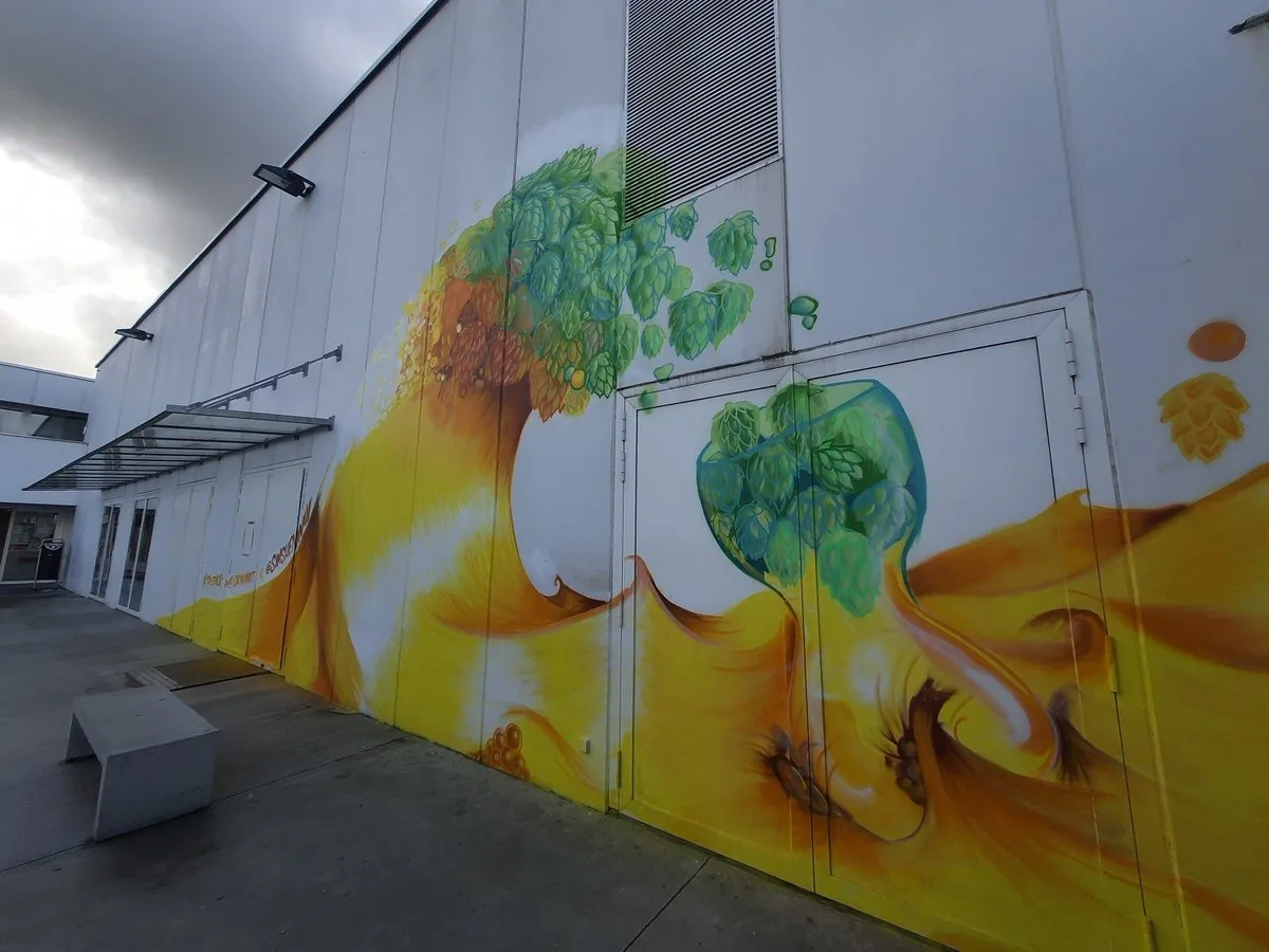 Large outdoor mural on a white building depicting a crashing wave of golden liquid and scattering green hops