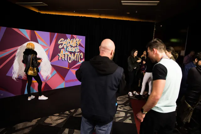 Woman poses at an event with a geometric mural reading SYDNEY GOES ATOMIC featuring a white hair silhouette
