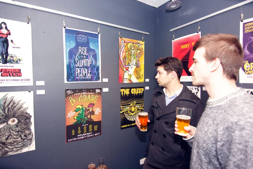 Two men holding beers while viewing illustrated B-movie style posters hanging on a dark gallery wall