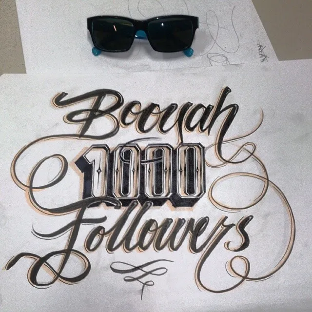Hand-drawn ink lettering on paper reading Booyah 1000 Followers, placed below a pair of black sunglasses