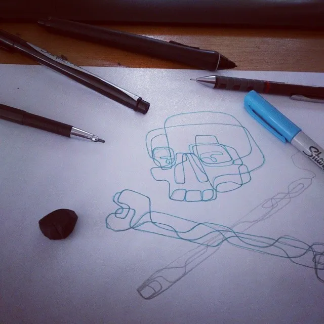 Blue line doodle of a stylized skull and crossbones on paper surrounded by pens and drawing tools