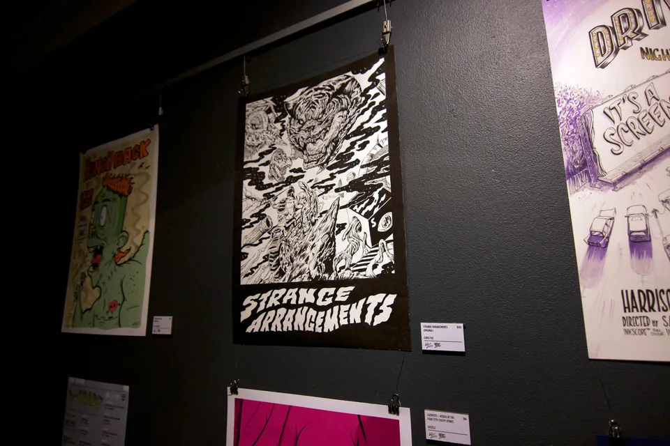 Black and white poster reading Strange Arrangements with a monstrous ink illustration hanging on a dark gallery wall