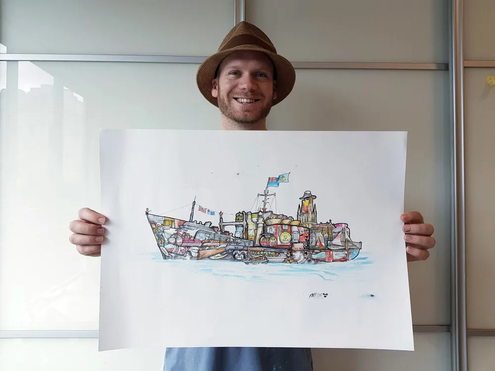 Smiling man in a hat holding a large illustration of a whimsical ship formed by clustered nautical objects and buildings