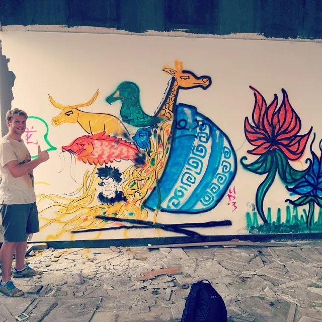 Man giving a thumbs up next to an in-progress mural of animals spilling from a noodle bowl and a large red flower