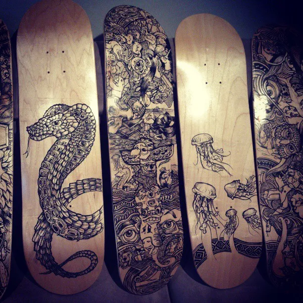 Wooden skateboard decks with black ink drawings of a patterned snake, dense totemic faces, and jellyfish