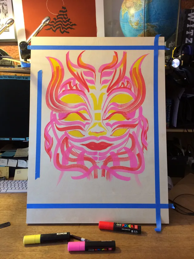 In-progress paint pen drawing on wood of a symmetrical, psychedelic face in flowing pink, red, and yellow lines