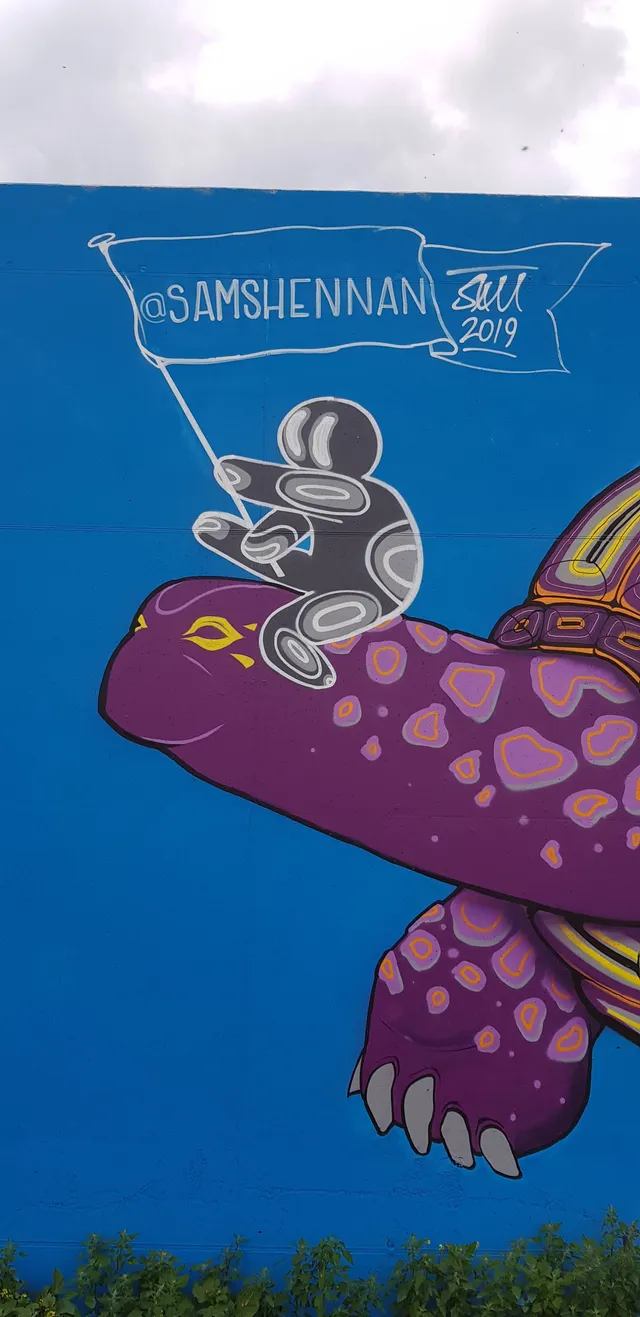 Mural of a stylized grey figure riding a large purple patterned tortoise, holding a flag on a blue wall
