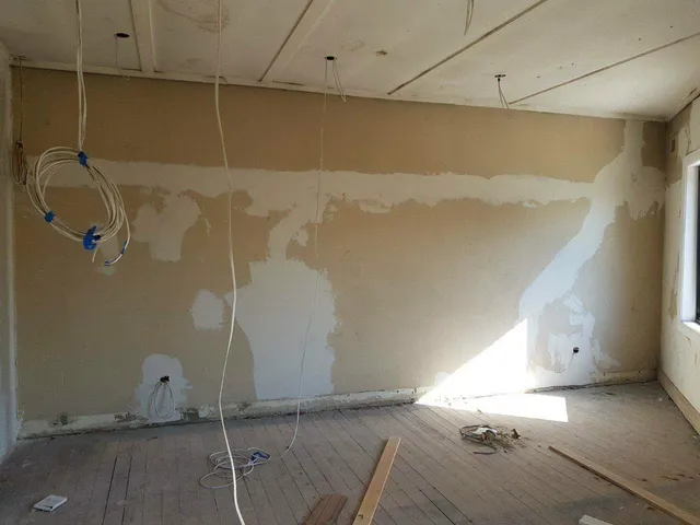 Unfinished room with a bare, patchy plaster wall, hanging electrical wires, and exposed wooden floorboards