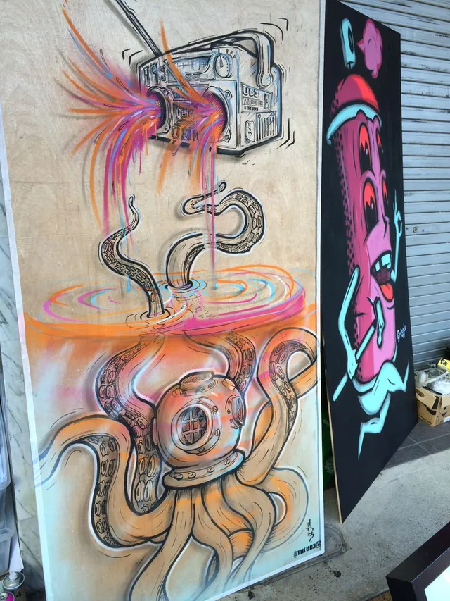 Murals on wood panels depicting a boombox spraying color over a diving-helmet octopus, beside a spray can character